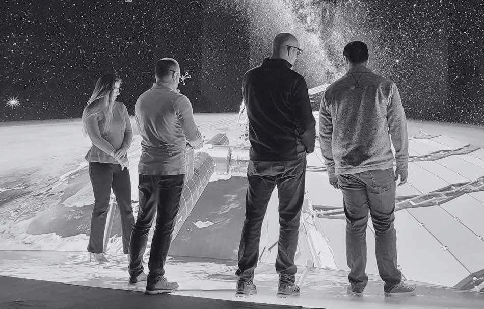 Black and white photo of 4 people standing in a large immersive visualization space with visuals of outer space displayed on the screens